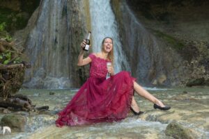 Sybille sitzend auf einem Stein vor dem Wasserfall im roten Abendkleid und schenkt sich ein Glas Sekt ein